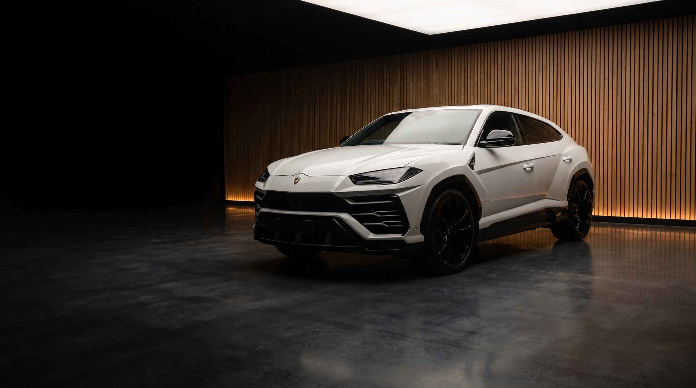 A white Lamborghini Urus alone in a dark showroom with an oak slatted wall